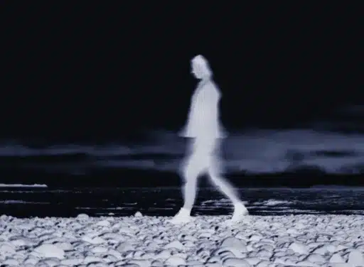 Glitchy, ghostly image of a women walking on a rocky beach with a black ocean in the background