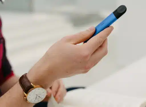 Person wearing gold round analog watch with brown leather strap holds a blue vape device
